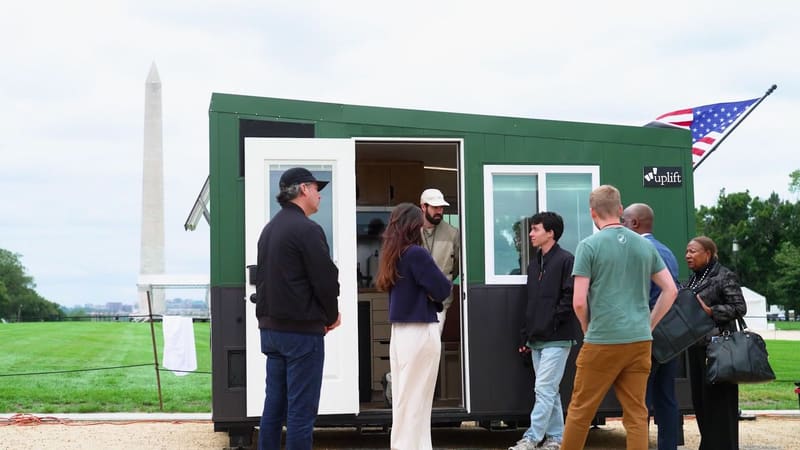 A green showcase home with its door open on the National Mall, visitors touring it, the Washington Monument and a U.S. flag behind.