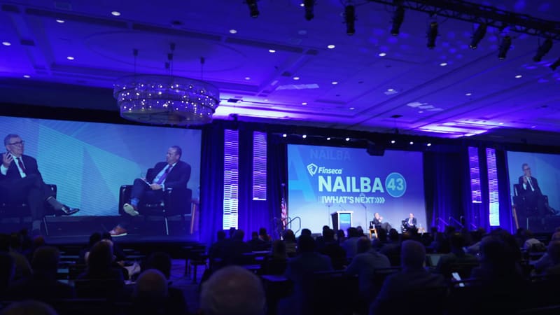 A wide view of a conference ballroom facing a stage with “NAILBA 43” on the screens.