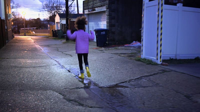 Seen from behind, a child in a purple jacket and yellow rain boots steps into a puddle in a wet alley at dusk, from Room to Breathe.